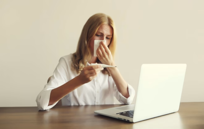 Erkältete Frau mit Taschentuch und Fieberthermometer am Laptop im Büro (Symbolbild), 06.10.2023. Foto: IMAGO / imagebroker (Veronika Korneva)